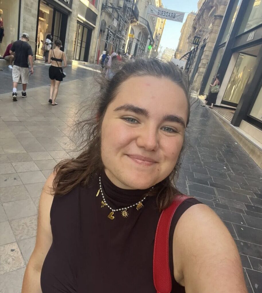 Jeune femme souriante prenant un selfie dans une rue commerçante, avec des magasins en arrière-plan